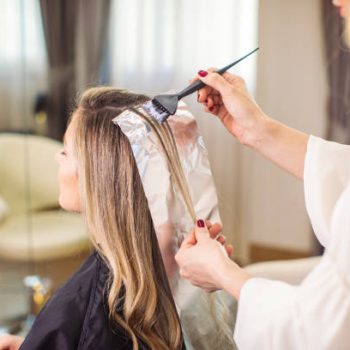 Blonde woman dyeing her hair at the salon. She is sitting and the hairdresser/hairstylist is putting hair color on her hair. This is regular hair care treatment in customers favorite salon.
